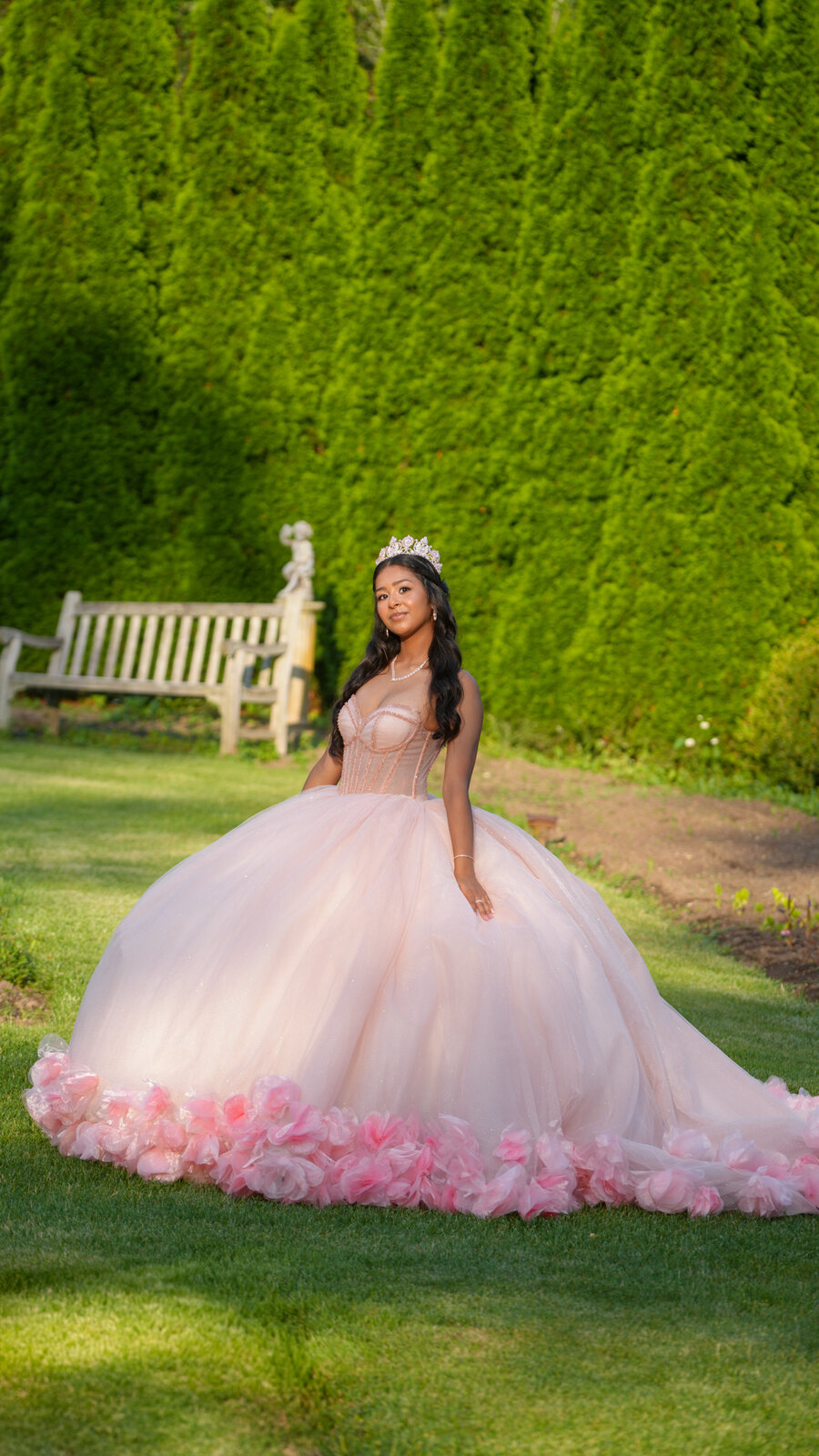 Quinceañera in a pink ball gown beside a tall hedge with a wooden bench behind her