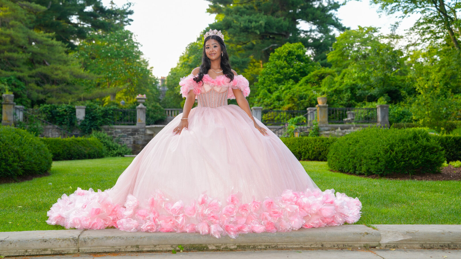 Quinceañera in a pink ball gown and tiara standing on a garden terrace