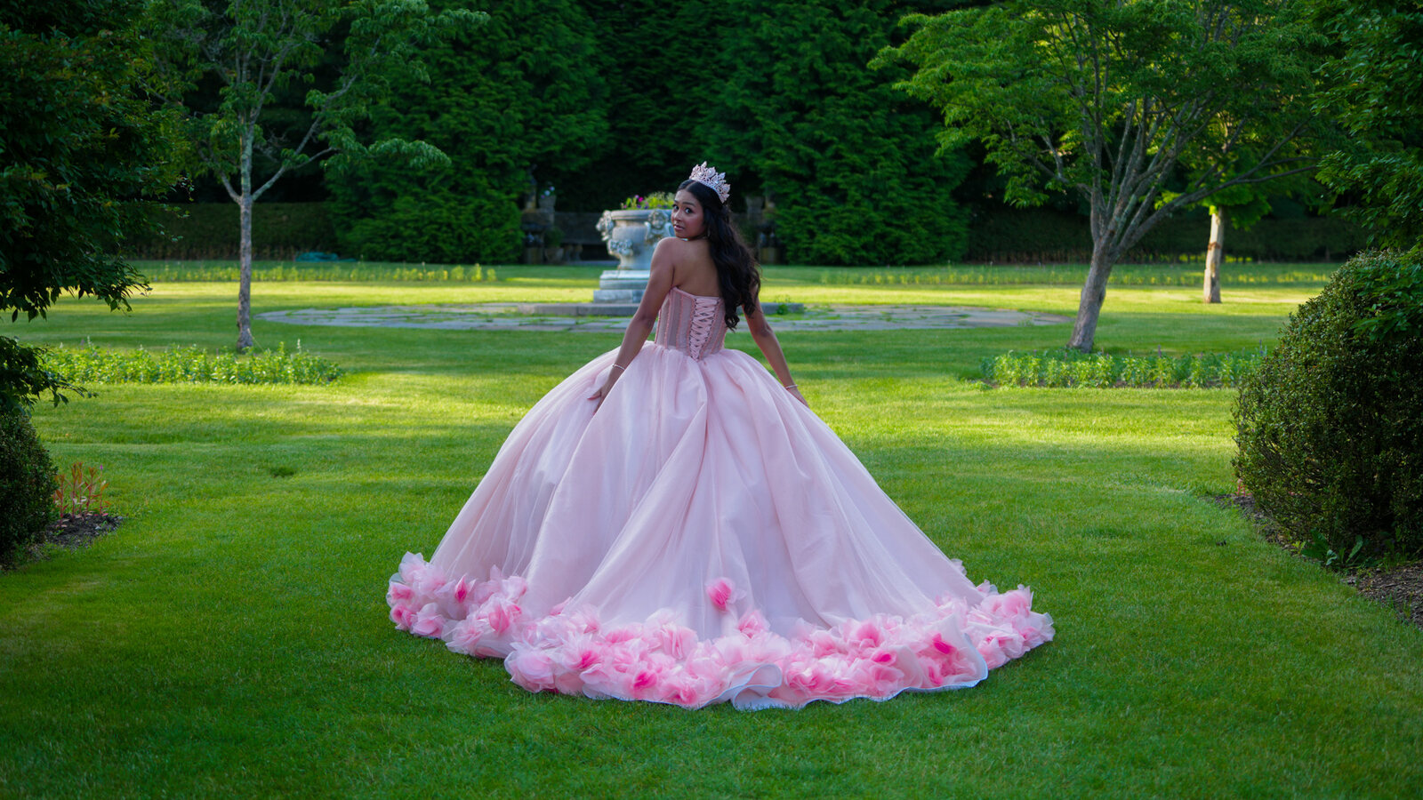 Quinceañera looking back over her shoulder with her pink dress spread on a garden lawn