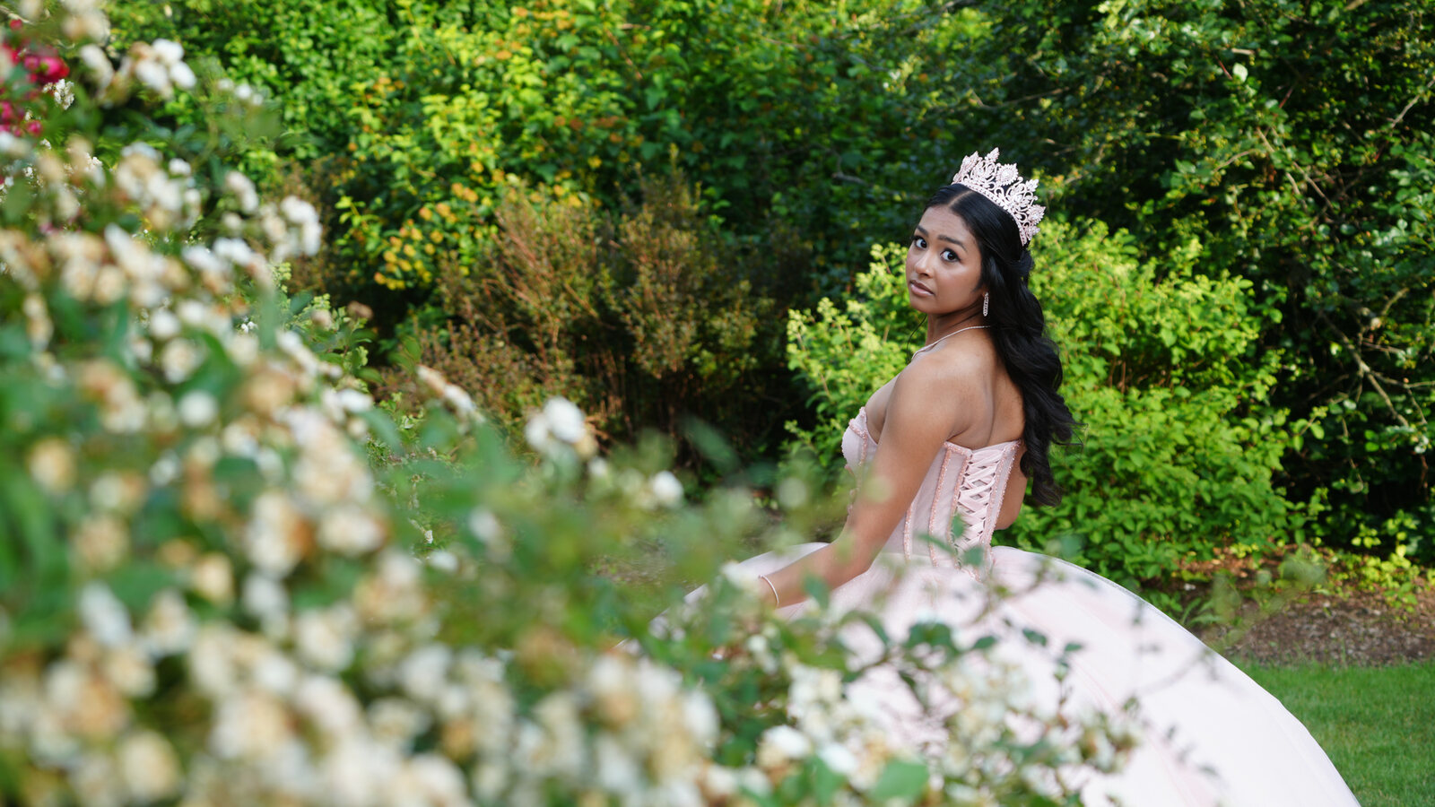 Quinceañera looking over her shoulder through white blossoms with her gown's lace-up back visible