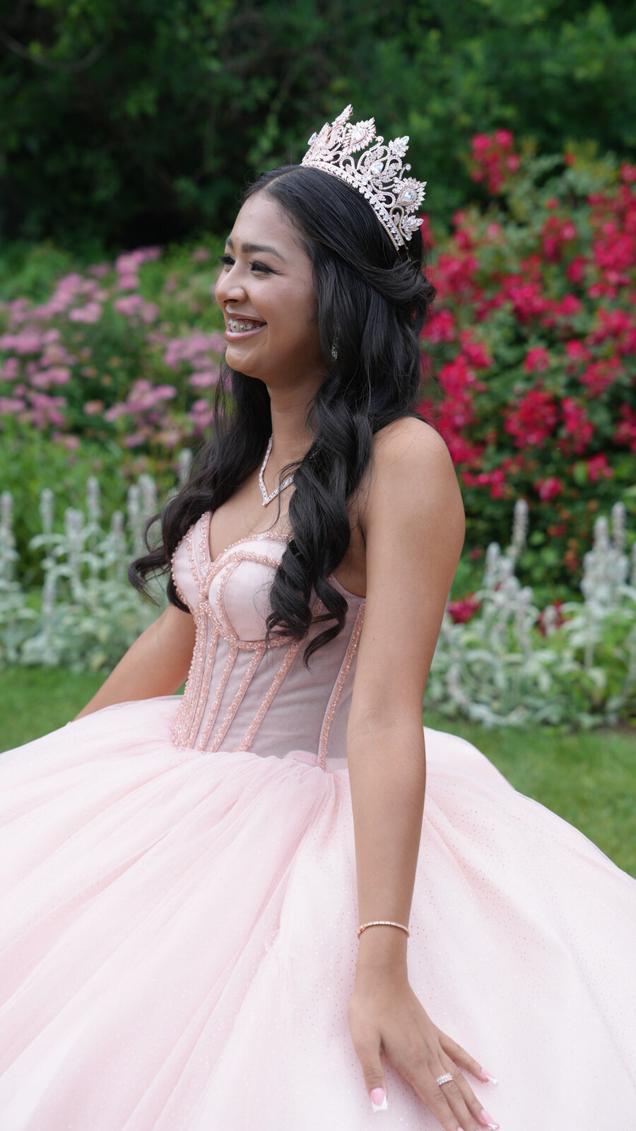 Quinceañera smiling in profile with a tiara and pink gown beside flowering roses