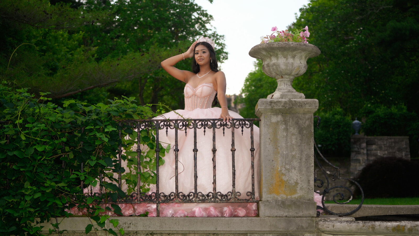 Quinceañera in a pink gown behind a wrought-iron railing beside a stone urn
