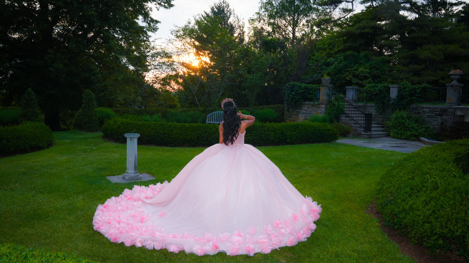 Back view of a quinceañera with her pink dress spread across a lawn at sunset
