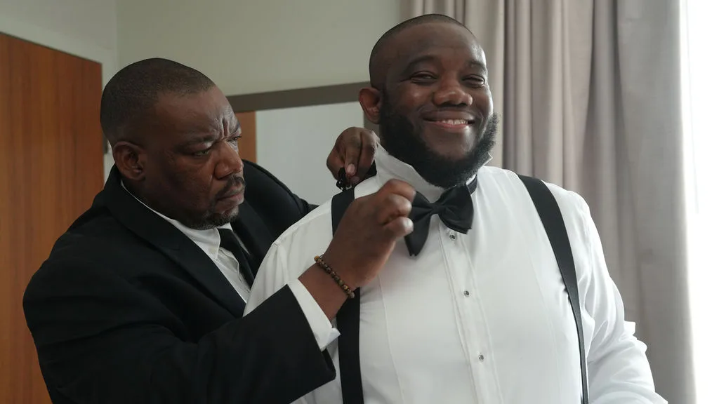 A loved one straightens Tre's bow tie in the groom's suite — generational wedding-morning moment, New Jersey