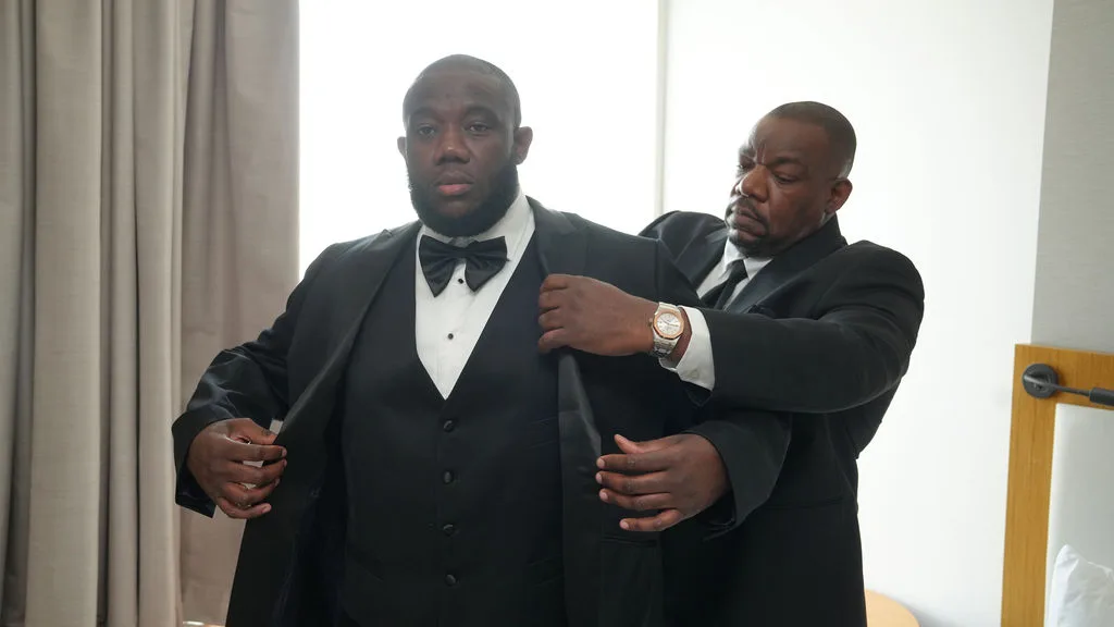 Tre is helped into his black tuxedo jacket in the hotel suite, wedding-morning candid, New Jersey