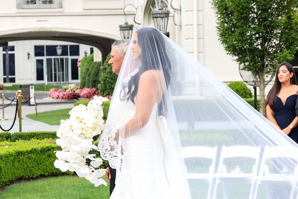 Bride carries cascading white orchid bouquet, cathedral veil trailing behind, processional at Park Chateau chapel, New Jersey
