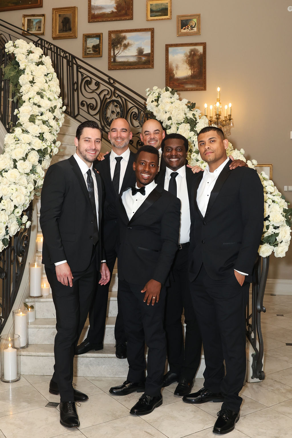 Groom and groomsmen in black tuxedos on the grand staircase at Park Chateau Estate, NJ, framed by white rose garlands