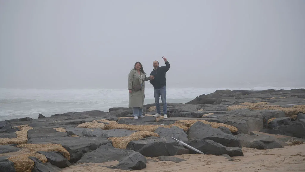 Just engaged — he flashes a peace sign, her hand in his, grinning on the jetty rocks, New Jersey shore