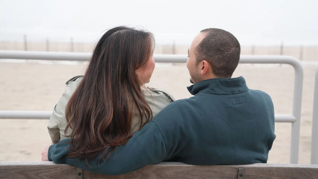 The newly engaged couple sits close on a boardwalk bench, looking at each other above the empty beach — New Jersey shore
